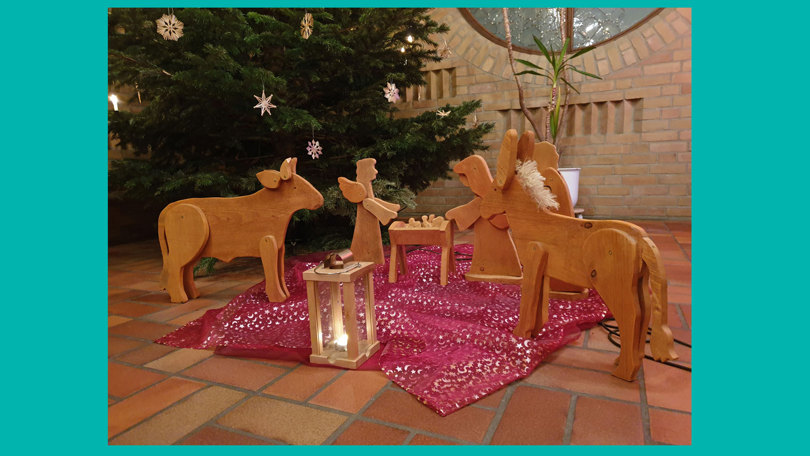 Krippenfiguren aus Holz, sie stehen auf einem rotgrundigen Tuch auf dem Boden im Kirchraum, dahinter ein mit Strohsternen geschm&uuml;ckter Weihnachtsbaum